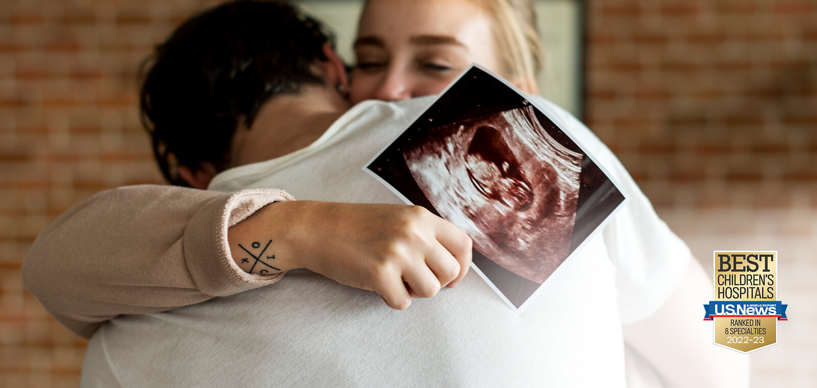 Two people embracing. One person holds image of sonogram depicting baby in a uterus. Awards badge for ranking as a best childrens hospital in 8 specialties. 