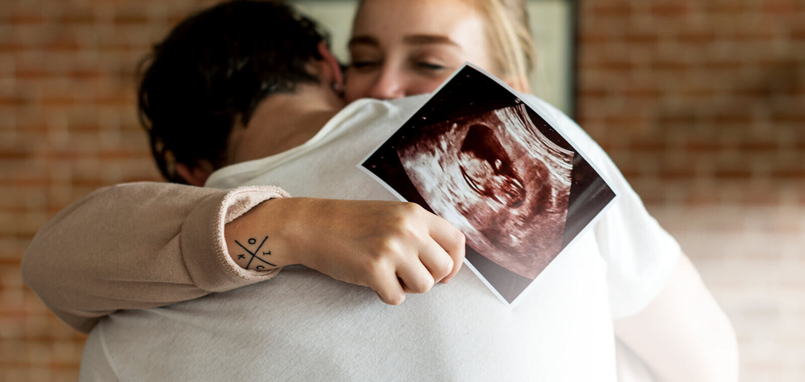 Two people embracing. One person holds image of sonogram depicting baby in a uterus.