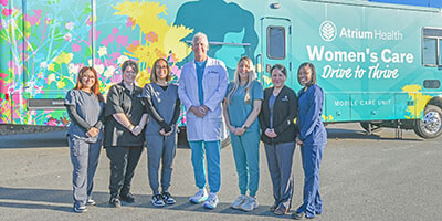 Eight women in scrubs of various colors and one man in a white lab coat stand near a colorful Mobile Care unit for a group photo.