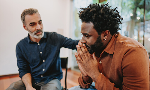A man with gray hair and a blue shirt comforts a man in an orange shirt.