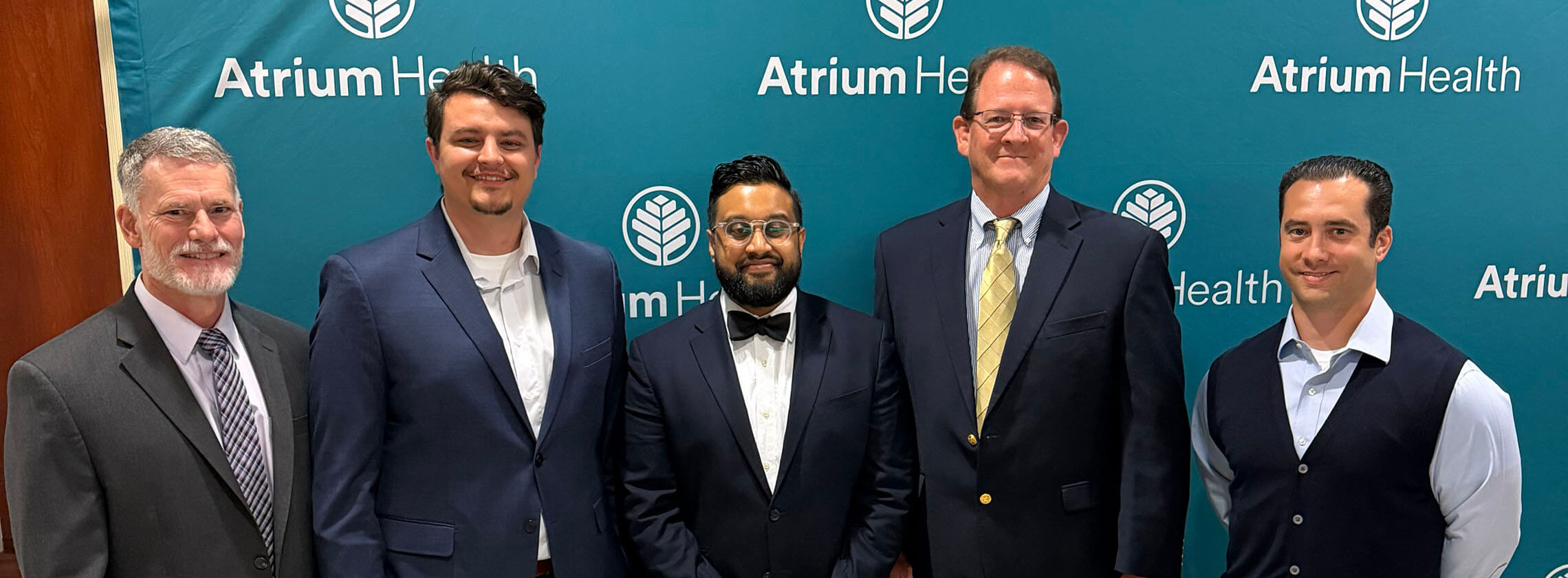 Four men in formal attire smiling in front of an Atrium Health teal backdrop.