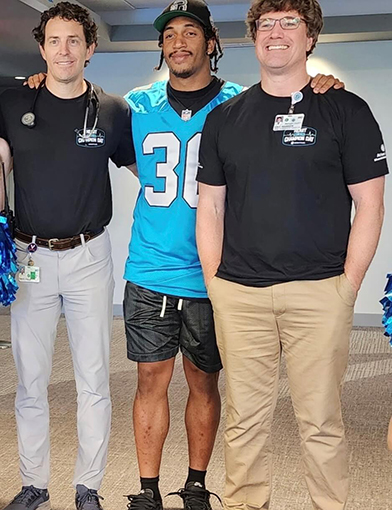 Two men standing with a football player in a light blue jersey.