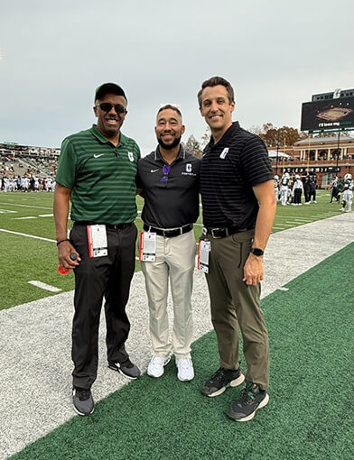Three men in polos and khakis standing on the sidelines of a football field.