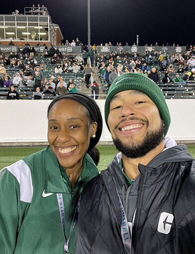 A young man in a black jacket and a young woman in a green jacket standing together on a football field.