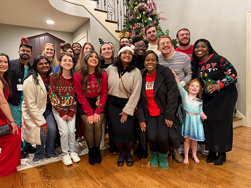 A group of men and women wearing holiday sweaters and clothes cathered by a Christmas tree.