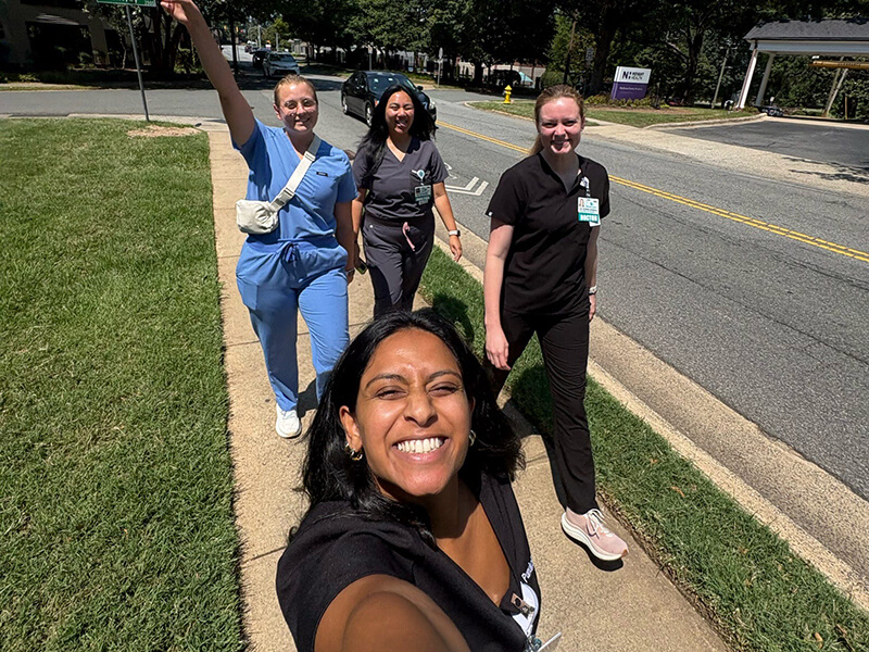 A woung woman with dark hair smiling while taking a selfie with three other women in scrubs.