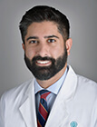 A man wearing a white lab coat sitting in front of a dark background.