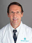 A headshot of a medical professional wearing a white lab coat and sitting in front of a dark background.