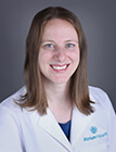 A woman with long hair, wearing a lab coat and smiling at the camera.