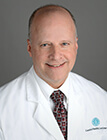 A man wearing a white lab coat, sitting in front of a dark background.