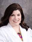 A medical professional wearing a lab coat and smiling at the camera in front of a dark background.