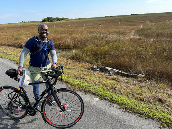 Jordan Mizell standing next to a bike near a crocodile on the side of a road in front of a field of tall grass.