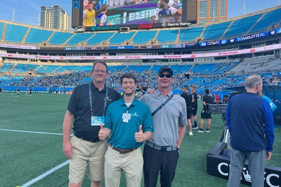 Three men standing together for a photo on the sidelines of a large football field and stadium.
