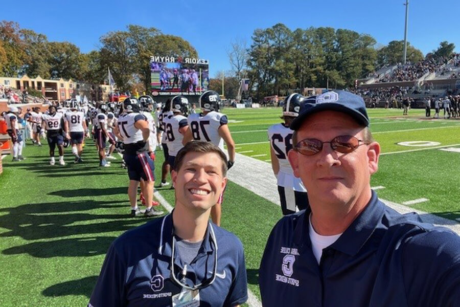 Two men wearing dark blue collared shirts standing on the sidelines of a football field.