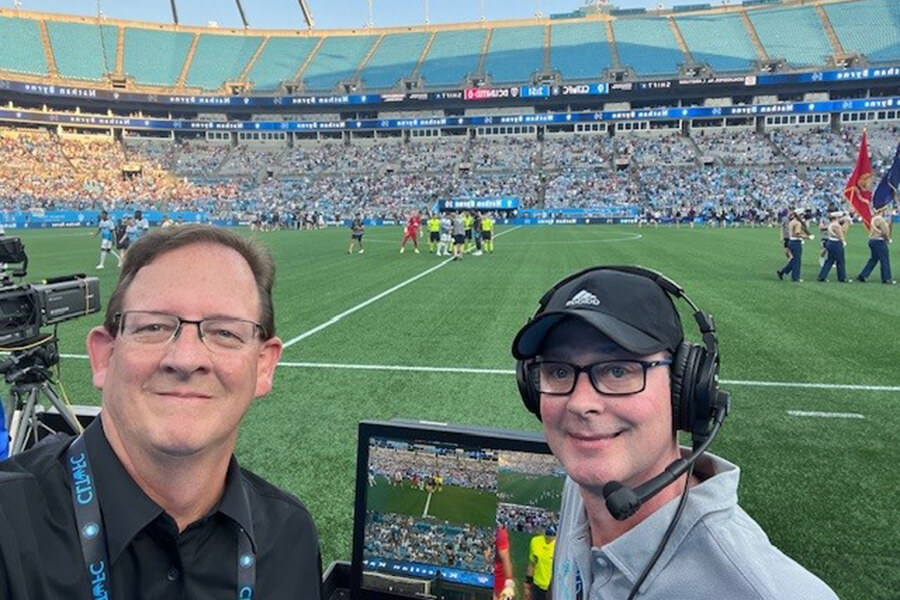 A man wearing a gray collared shirt and a large headset standing next to a small video screen alongside a man in a black shirt.