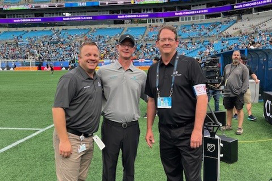 Three men in gray and black shirts standing together on the sideline of a sports field.