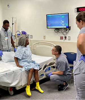 A woman in a hospital gown sits on a bed while talking to nurses and doctors.