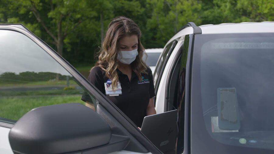 A woman wearing medical scrubs and a face mask standing next to a car.