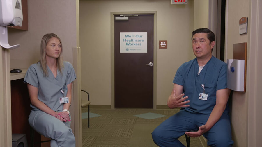 Two medical professionals wearing scrubs, sitting on stools and talking.