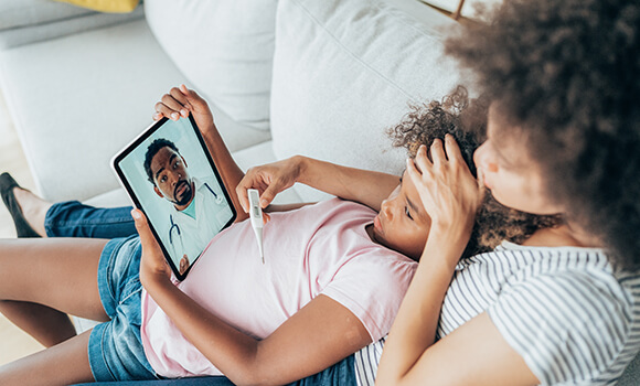A woman sitting on a couch with a child in her lap consulting virtually with a doctor.