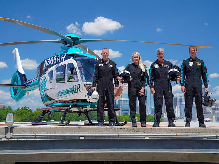 A group of four pilots in flight suits stand on a helipad next to a helicopter.