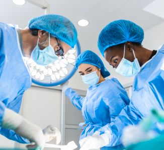 Three doctors wearing blue scrubs and blue masks preforming surgery.