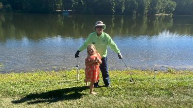 Tom wearing green jacket holding fishing poles and fish in front of water on grass with young girl in pink.
