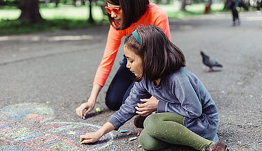 A young adult and a child in long sleeve shirts and pants drawing on asphalt with chalk.