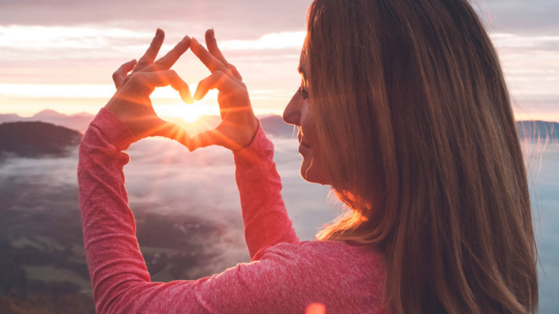 Woman on a mountaintop making heart with hands with sun in the middle