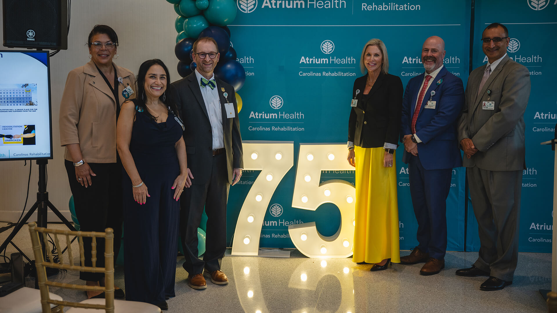 A group of men and women in formal attire smiling in a group next to large light up numbers showing the number 75.