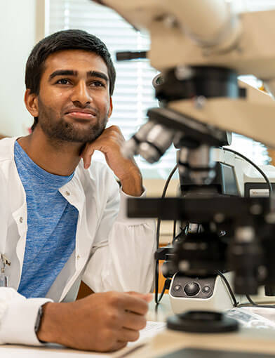 A man with dark hair wearing a white lab coat at his desk with a microscope.