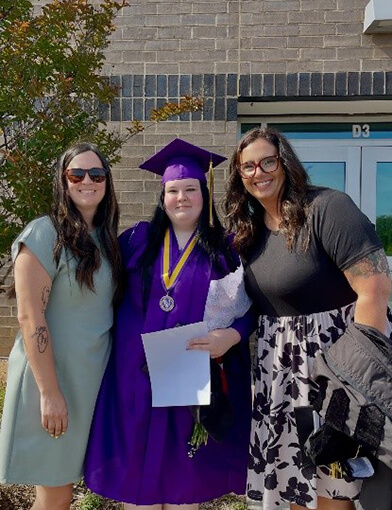 A young woman in a blue cap and gown standing in between two women in dresses.