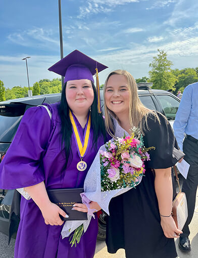 A young woman in a blue cap and gown standing next to another young woman in a black dress.