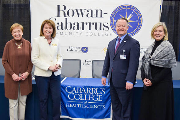 Faculty and administrative members in formal attire standing in front of a Rowan-Cabarrus banner and a table.