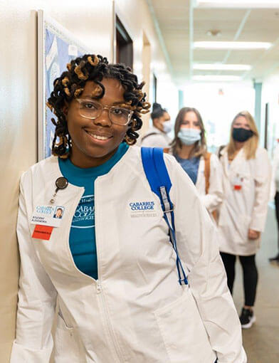 An African American Nurse in a white coat smiling while standing in a hallway.