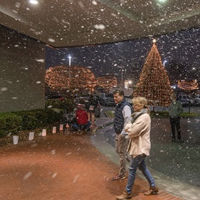A couple walking through snow flurries with a decorated tree in the background.