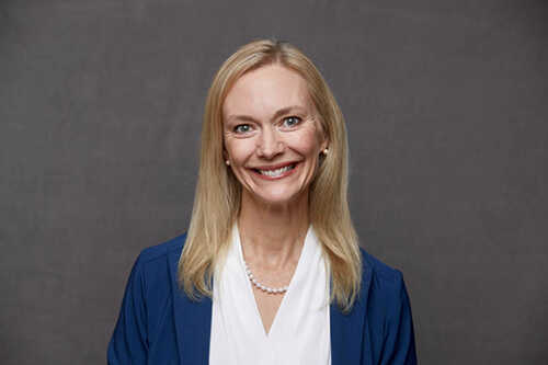 A women with long blonde hair wearing a white shirt and blue jacket smiling at the camera.