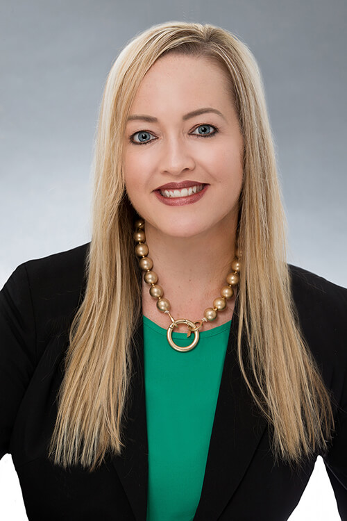 A young woman with long blonde hair wearing a green shirt and black jacket smiling at the camera.