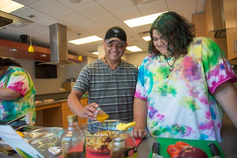 An older person wearing a hat pours dressing over a dish while standing next to a younger person wearing a tie-dye shirt.