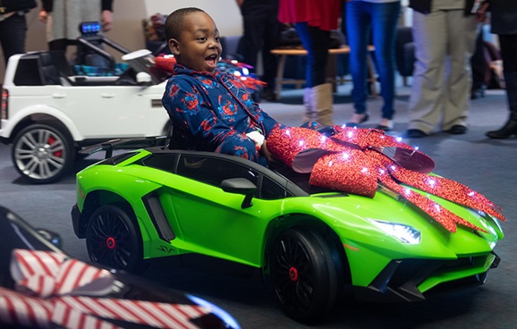A young Black boy in pajamas laughs as he drives a bright green miniature motorized car with lights and a holiday bow on the hood inside Brenner Children's