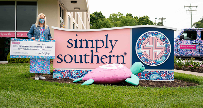 A blonde woman in a face mask and patterned dress holds an oversize check while standing next to the corporate sign for Simply Southern. A turquoise and pink giant turtle sculpture sits on the ground in front of the sign