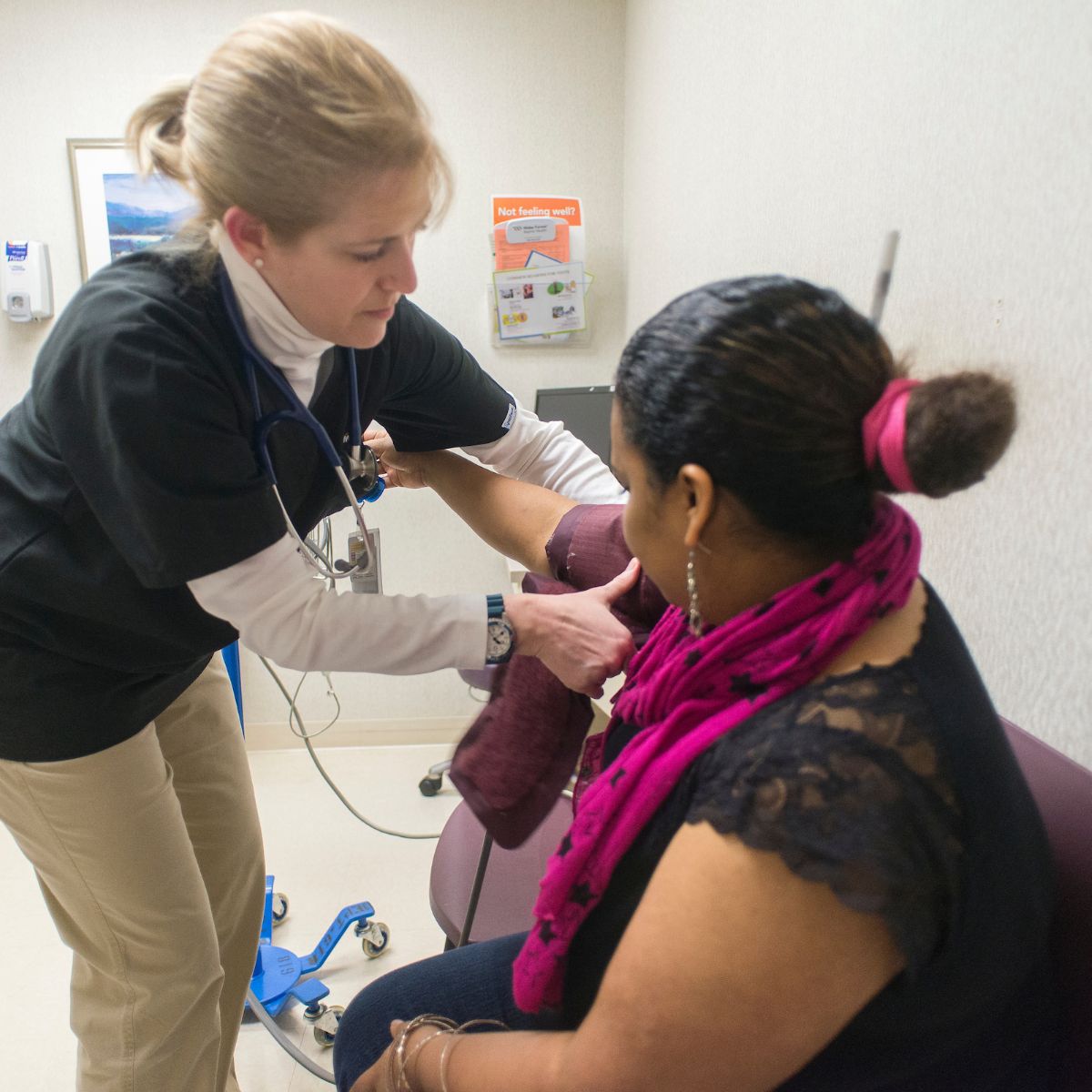 Nurse with blonde hair taking blood pressure of woman with pink scarf who is sitting down.