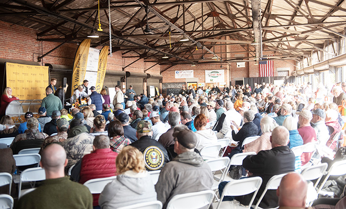 Large crowd of people in cold weather clothing sit in white chairs in an open-air train depot listening to a speaker/auctioneer