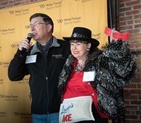 A man in dark sweater speaks into a microphone as a woman in a fur coat, black hat and red shirt holds up raffle tickets