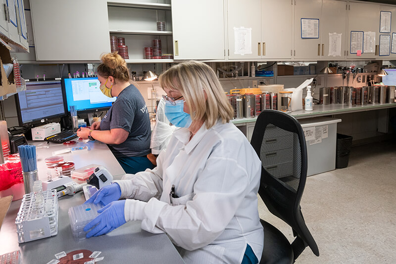 Two women at lab desk both wearing mask doing work