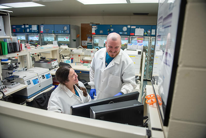 A man in a white labcoat stands while pointing at a monitor giving instructions to a woman in a white labcoat.