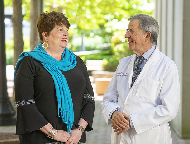 A woman and older man in white coat stand outside and smile at each other