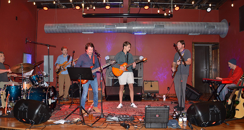 Five men and one male teen play various instruments on stage during a fundraiser concert