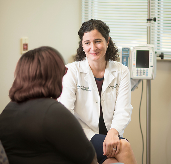 White female with dark hair wearing white coat smiles at dark-haired female patient sitting in chair