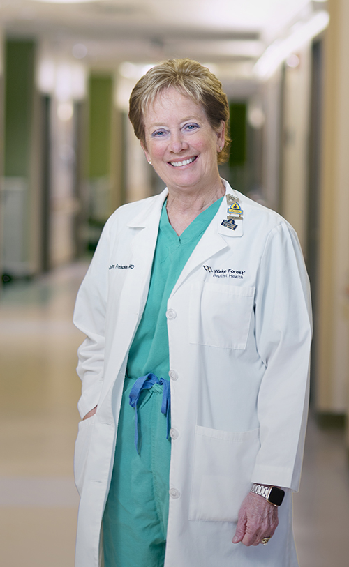 White woman in green scrubs and white coat smiles and stands in hallway